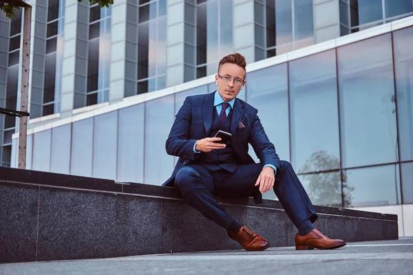 一个穿着优雅西装的自信时髦的商人的肖像手持智能手机, 看着相机, 同时坐在户外对摩天大楼背景. — 图库照片