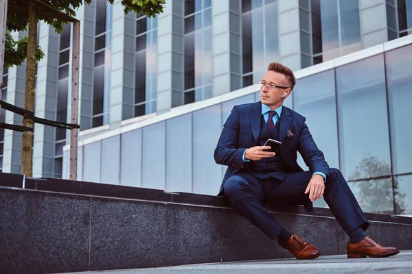 Portrait of a confident stylish businessman dressed in an elegant suit holds a smartphone and looking away while sitting outdoors against a skyscraper background. — Stock Photo, Image