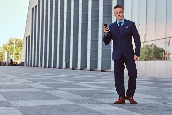 Hela kroppen porträtt av en säker affärsman klädd i en elegant kostym rymmer en telefon stående mot stadsbilden bakgrund. — Stockfoto