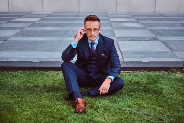 Hombre de negocios pensativo vestido con traje elegante sentado en un césped verde contra el fondo del paisaje urbano . —  Fotos de Stock