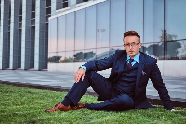 Hombre de negocios confiado vestido con traje elegante sentado en un césped verde contra el fondo del paisaje urbano . —  Fotos de Stock
