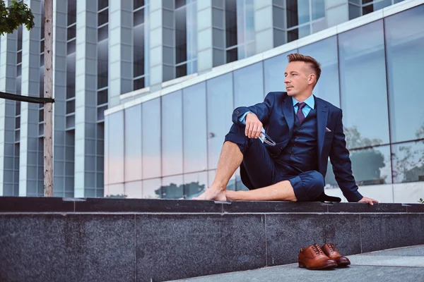 Confident businessman dressed in an elegant suit holds his glasses while sitting barefoot against cityscape background. — Stock Photo, Image