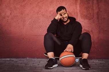 Afrikalı-Amerikalı basketbol ile bir kaykay üzerinde oturan ve duvara yaslanmış bir siyah hoodie ve spor şort giymiş adam sakallı.