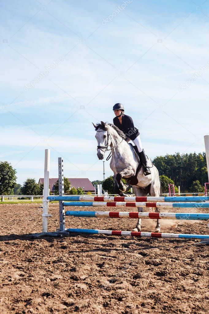 Female jockey on dapple gray horse jumping over hurdle in the open arena.