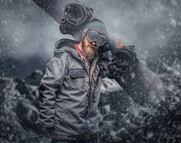 残酷的红发滑雪板与一个完整的胡子在冬季帽子和防护眼镜穿着滑雪板大衣举行滑雪板反对山的背景 — 图库照片