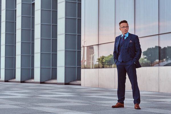 Confident businessman dressed in an elegant suit looking away and holds hands in pockets while standing outdoors against cityscape background.