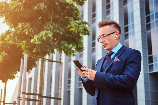 Retrato de um empresário elegante e confiante vestido com um terno elegante usando um smartphone enquanto está ao ar livre contra o fundo do arranha-céu . — Fotografia de Stock