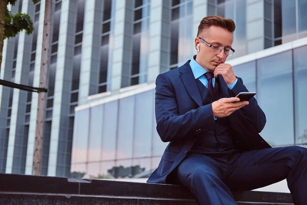 Retrato de un hombre de negocios con estilo y confianza vestido con un traje elegante usando un teléfono inteligente mientras está sentado al aire libre contra el fondo del rascacielos . —  Fotos de Stock