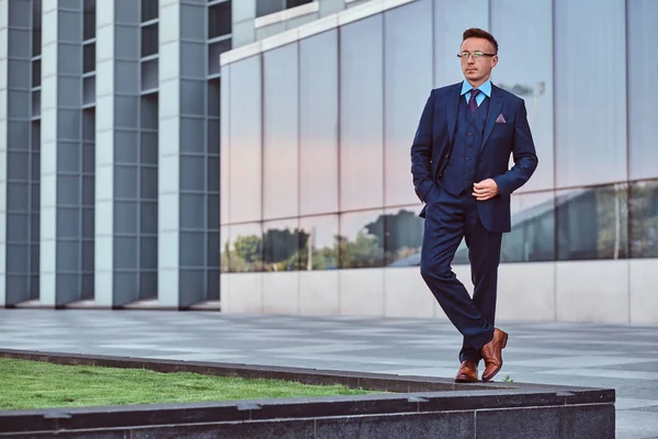 Full body portrait of a confident man in an elegant suit posing with hand in pocket against skyscraper background.