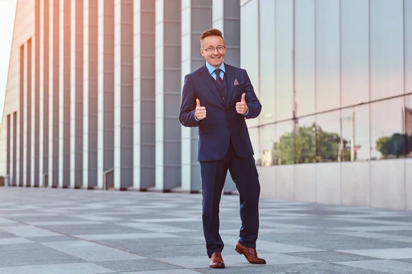 Joyful säker affärsman klädd i en elegant kostym visar gest cool medan stående utomhus mot stadsbilden bakgrund. — Stockfoto