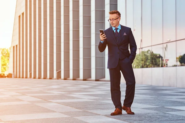 Hela kroppen porträtt av en säker affärsman klädd i elegant kostym använder en telefon stående mot stadsbilden bakgrund. — Stockfoto