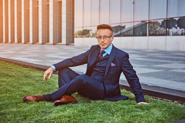 Empresário confiante vestido em terno elegante sentado em um gramado verde contra o fundo da paisagem urbana . — Fotografia de Stock