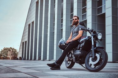 Gri tişört ve siyah pantolon acımasız bir sakallı erkek portresi bir gökdelen karşı özel yapım retro motosiklet üzerinde oturan bir kask tutar