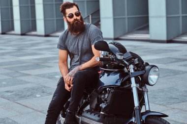 Gri tişört ve siyah pantolon bir gökdelen karşı özel yapım retro motosiklet üzerinde oturan bir acımasız sakallı bisikletçinin güneş gözlüğü portresi giymiş