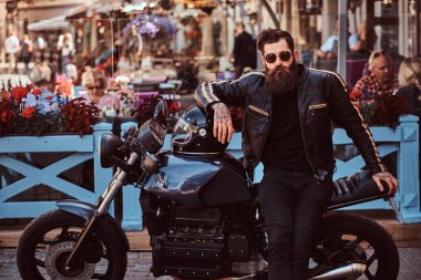 Güneş gözlüklü bir kafe Teras yakınındaki özel yapım retro motosiklet üzerinde oturan bir siyah deri ceket giymiş bir şık şık motorcu portresi.