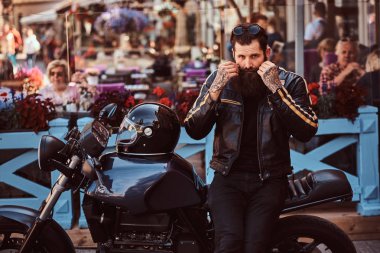 Bir siyah deri ceket giymiş bir şık şık motorcu portresi düzeltme bıyık bir kafe Teras yakınındaki özel yapım retro motosiklet üzerinde otururken.