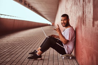 Neşeli Afrikalı-Amerikalı sakallı adam bir beyaz giyinmiş gömlek ve spor şort tutan bir dizüstü bilgisayarda bir köprü altında bir kaykay üzerinde otururken bir kameraya bakıyor.