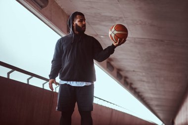 Köprü altında bir footway üzerinde bir basketbol duran siyah bir hoodie ve spor şort giymiş esmer bir adam portresi tutar.