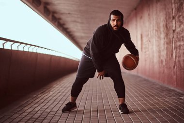 Bir kameraya bakarak adam koyu tenli bir footway bir köprü altında basketbol oynamak siyah bir hoodie ve spor şort giymiş.