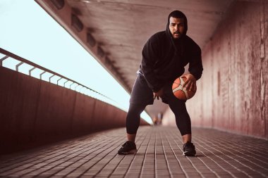 Bir kameraya bakarak adam koyu tenli bir footway bir köprü altında basketbol oynamak siyah bir hoodie ve spor şort giymiş.