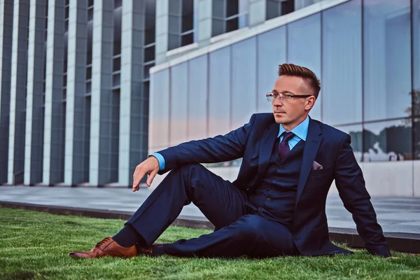 Hombre de negocios confiado vestido con traje elegante sentado en un césped verde contra el fondo del paisaje urbano . —  Fotos de Stock