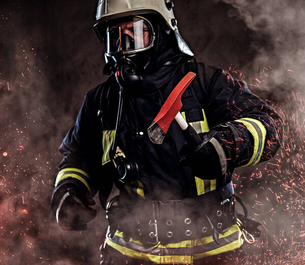 A firefighter dressed in a uniform and an oxygen mask holds a red axe standing in fire sparks and smoke over a dark background