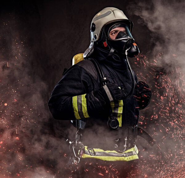 Портрет пожарного в форме и кислородная маска, стоящая в огненных искрах и дыма на темном фоне
.