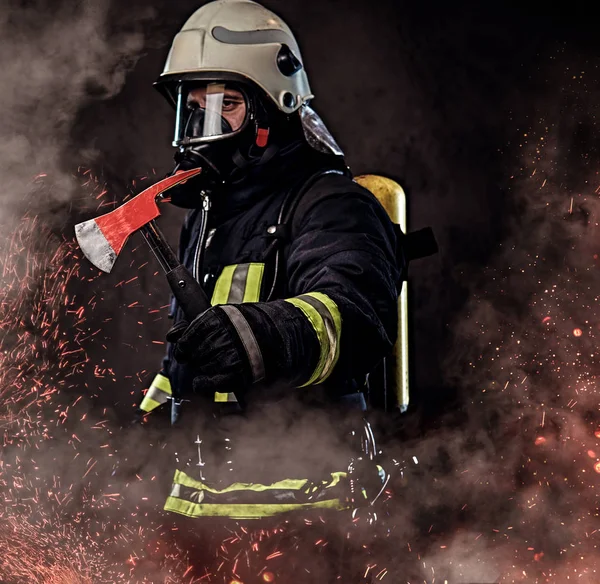 一名身穿制服和氧气面罩的消防员手持一把红色斧头站在火的火花和烟雾在黑暗的背景 — 图库照片