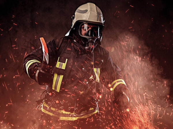 A firefighter dressed in a uniform and an oxygen mask holds a red axe standing in fire sparks and smoke over a dark background