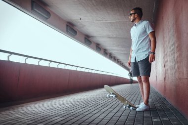 Uzak seyir yakışıklı şık patenci adam beyaz bir gömlek ve şort, köprünün altından bir patika üzerinde duran bir duvara yaslanmış giymiş güneş gözlüğü.