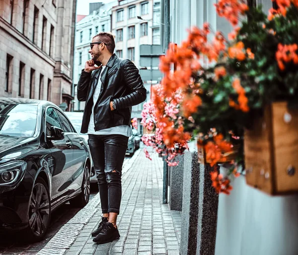 时尚的家伙在太阳镜身着黑色的皮革夹克和牛仔裤谈话由智能手机 而站在豪华车附近的旧欧洲街 — 图库照片