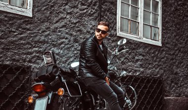 Güneş gözlüklü bir siyah deri ceket ve retro motosikleti eski Avrupa sokakta oturan kot giymiş şık genç motorcu.