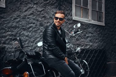 Güneş gözlüklü bir siyah deri ceket ve retro motosikleti eski Avrupa sokakta oturan kot giymiş şık genç motorcu.