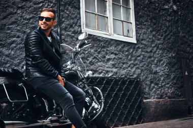 Güneş gözlüklü bir siyah deri ceket ve retro motosikleti eski Avrupa sokakta oturan kot giymiş şık genç motorcu.