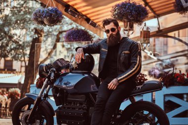 Güneş gözlüklü bir kafe Teras yakınındaki özel yapım retro motosiklet üzerinde oturan bir siyah deri ceket giymiş bir şık şık motorcu portresi.