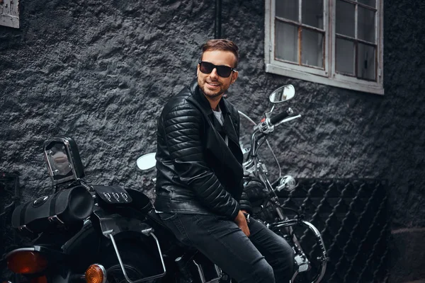 时尚的年轻车手在太阳镜身着黑色的皮革夹克和牛仔裤坐在他的复古摩托车在旧欧洲街 — 图库照片