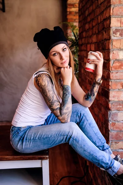 Chica hipster tatuada pensativa vestida con camiseta blanca y sombrero sostiene el café para llevar mientras está sentado en la mesa en una habitación con interior loft . — Foto de Stock
