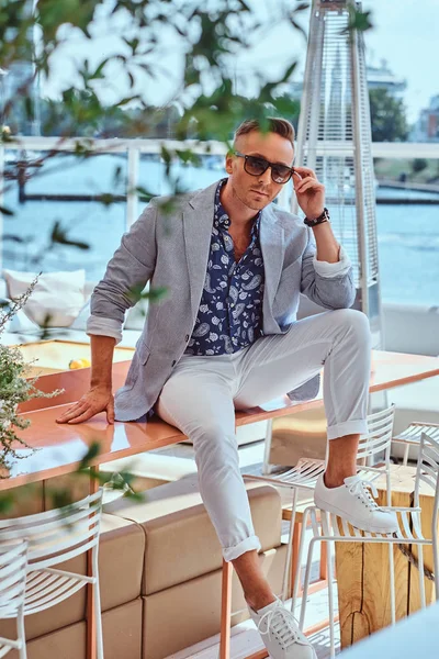 成功时尚男士穿着现代高雅的衣服坐在户外咖啡馆的桌子上, 在城市码头的背景下. — 图库照片