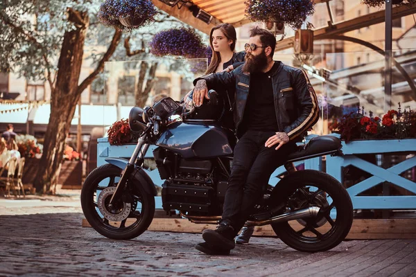 Casal hipster atraente - macho brutal barbudo em óculos de sol vestidos com uma jaqueta de couro preto e sua jovem menina sensual de pé perto, posando contra o terraço do café . — Fotografia de Stock