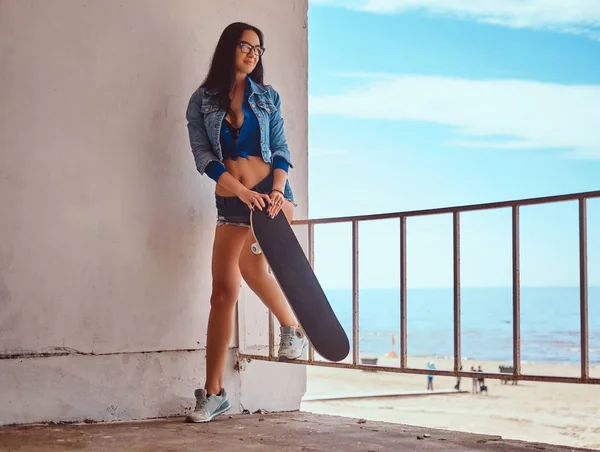 ショート パンツやジーンズのジャケットを身に着けている魅惑的なホット ブルネットの少女は海の海岸に対してガードレール頼りに立ちながらスケート ボードを保持します — ストック写真
