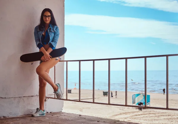 ショート パンツやジーンズのジャケットを身に着けている魅惑的なホット ブルネットの少女は海の海岸に対してガードレール頼りに立ちながらスケート ボードを保持します — ストック写真