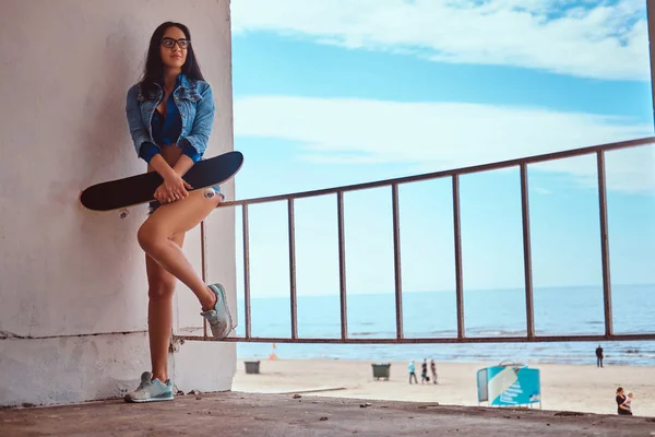 Baştan çıkarıcı seksi esmer kız kısa şort ve kot ceket giyen bir kaykay bir Otokorkuluk bir yalı karşı sırtını dayayacak ayakta iken tutar.