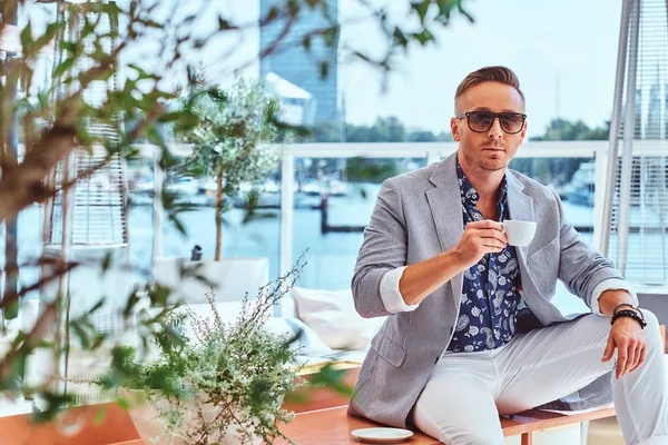 Selbstbewusster erfolgreicher Mann mit stylischem Haar in moderner eleganter Kleidung hält eine Tasse Kaffee in der Hand, während er auf einem Tisch im Café im Freien vor dem Hintergrund der Stadtmole sitzt. — Stockfoto