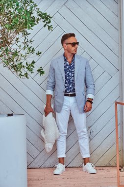 Modern şık giysiler giymiş güneş gözlüğü moda adam beyaz bir ahşap duvar karşı duran bir balık heykel tutar.