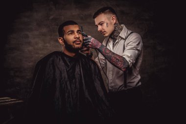 Eski moda profesyonel dövmeli Kuaför saç tıraşına bir düzeltici ve tarak kullanarak bir Afro-Amerikan istemciye kuramıyor. Karanlık dokulu arka plan üzerinde izole