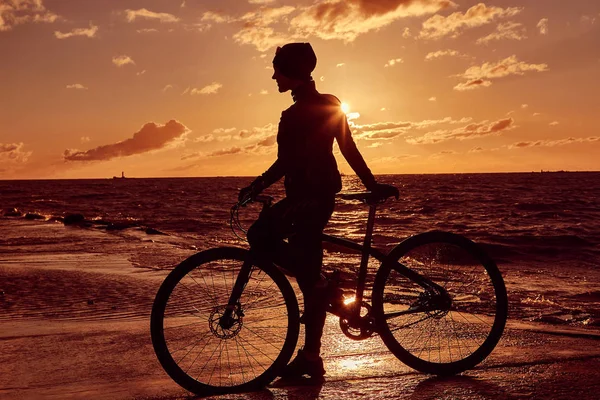 Silhouette of a girl with a bicycle on a sea coast on a sunset background. — Stock Photo, Image