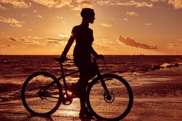 Female cyclist standing with her bicycle and enjoying the sunset on the sea coast. — Stock Photo, Image