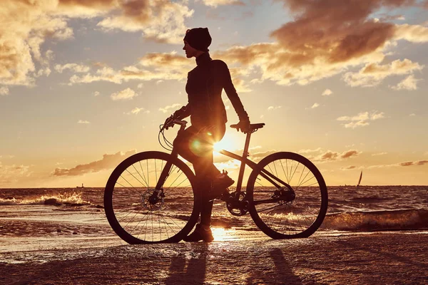 Female cyclist standing with her bicycle and enjoying the sunset on the sea coast. — Stock Photo, Image