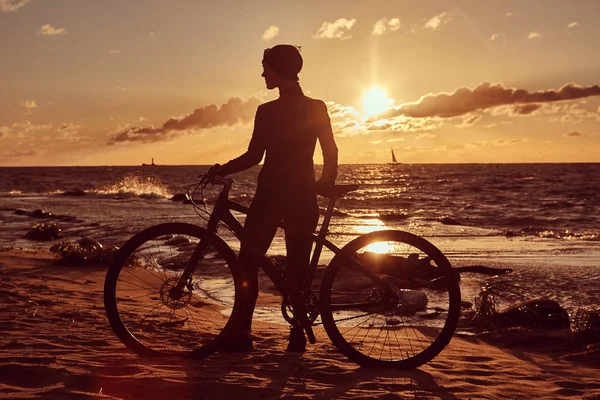 Female cyclist standing with her bicycle and enjoying the sunset on the sea coast. — Stock Photo, Image