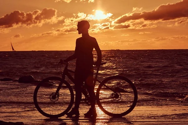 彼女は自転車の側に立って、海の海岸に夕日を楽しんで女性サイクリスト. — ストック写真